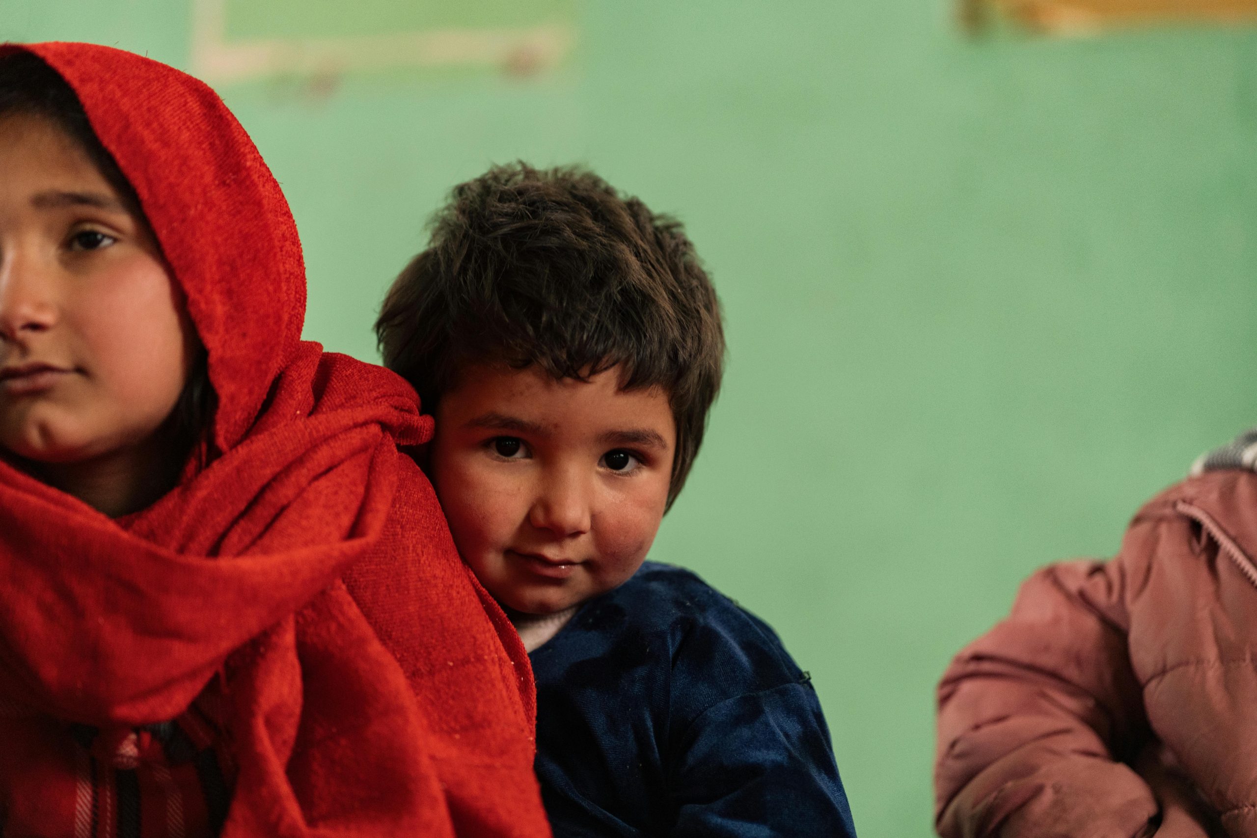 Two Afghan children in traditional clothing, captured in a candid moment indoors.