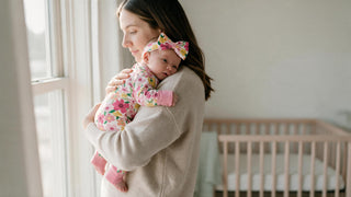 Mother holding a newborn in a cream footed sleeper by a window, illustrating newborn versus 0-3 month sizing