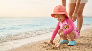 Toddler in a long-sleeve rash suit, tie-on sun hat and water shoes digging in wet sand at the shoreline in morning light